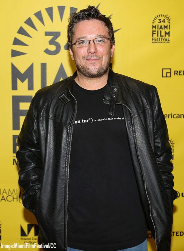 Filmmaker, Bily Corben, stands in front of a bright yellow Miami Film Festival backdrop wearing a black leather jacket, jeans, and a black T-shirt that reads “one who tells stories.” He has short dark hair, glasses, and a relaxed smile. The image credit reads: Image: MiamiFilmFestival/CC.