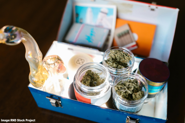 An open metal box containing cannabis jars filled with buds, a glass smoking pipe, rolling papers, and other accessories for smoking. The contents are neatly organized and brightly lit. Text in the bottom left corner reads “Image: RND Stock Project.