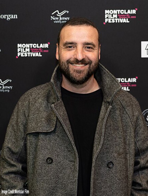 Actor David Krumholtz stands smiling in front of a black Montclair Film Festival backdrop with logos for JPMorgan and New Jersey Visit. He wears a dark gray wool coat over a black shirt. The lighting highlights his face against the festival’s red and white text. Image Credit: Montclair Film.