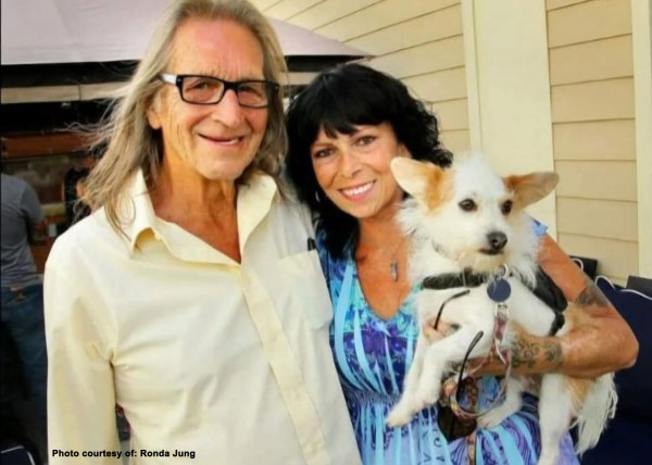 George Jung smiles in a pale yellow shirt beside Ronda Jung, who is holding a small white dog with tan ears. They are standing outside near a beige house with a patio umbrella visible in the background. The photo is credited to Ronda Jung.