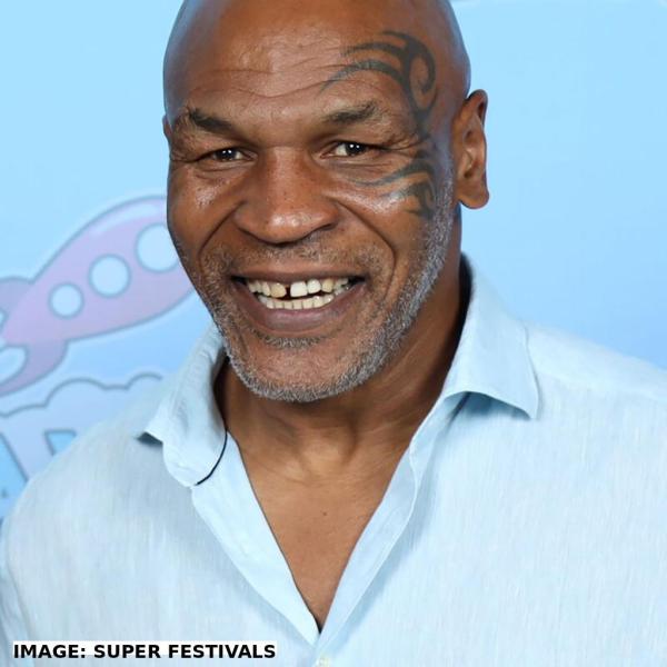 Former heavyweight boxing champion Mike Tyson smiles during a photo op at GalaxyCon Austin 2023. He wears a light blue shirt and sports his signature face tattoo and a gray beard. The background features playful cartoon graphics. Image credit: Super Festivals.