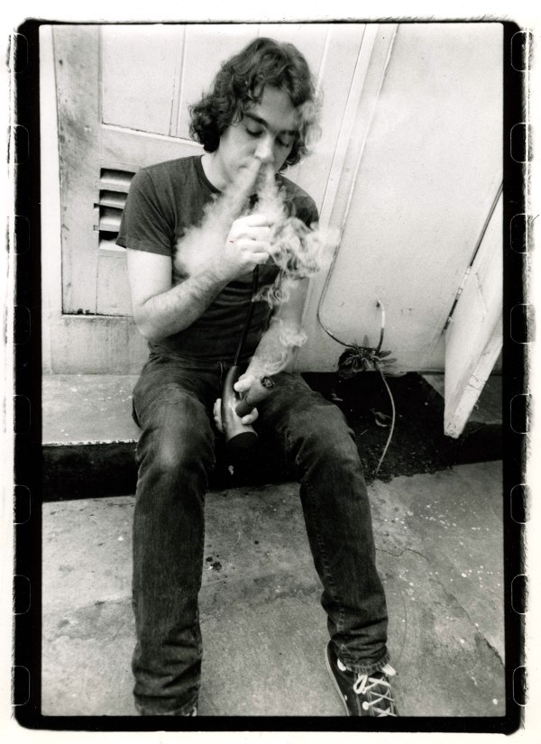 Former High Times Magazine Editor-in-Chief Chris Simunek sits on a concrete step, smoking from a pipe as smoke swirls around his face in a black-and-white photograph.