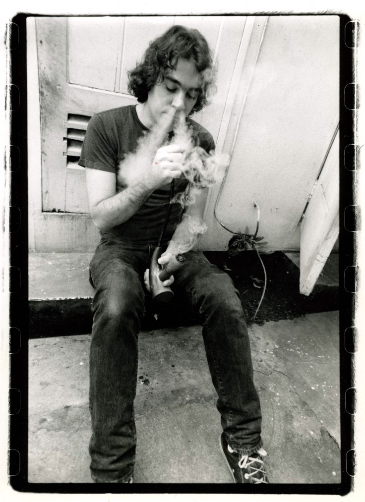 Former High Times Magazine Editor-in-Chief Chris Simunek sits on a concrete step, smoking from a pipe as smoke swirls around his face in a black-and-white photograph.