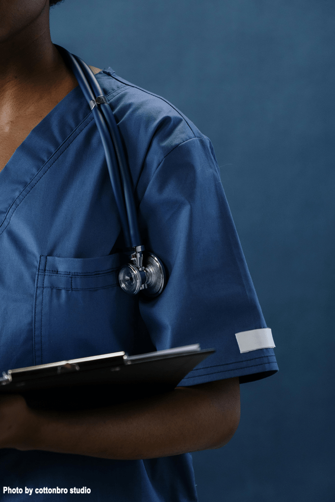 A close-up of a healthcare professional in blue scrubs holding a clipboard, with a stethoscope draped around their neck against a dark blue background.
