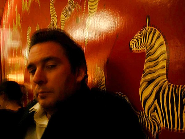 Chris Simunek sits indoors in front of a deep red wall decorated with painted zebras. He has short dark hair and wears a dark jacket over a white shirt. The lighting is dim and warm, giving the scene a moody, artistic tone. A watermark in the corner reads “Image by: Rebekah Harris