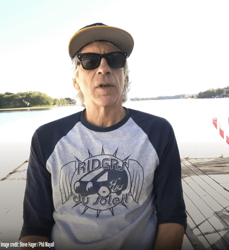 Steven Hager wearing sunglasses, a baseball cap, and a “Riders du Soleil” shirt, sitting on a dock by a calm lake with trees and sky in the background.