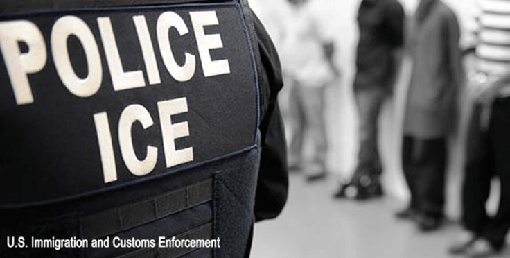 Close-up of a U.S. Immigration and Customs Enforcement (ICE) officer’s vest with the word ‘POLICE’ visible, while a blurred line of detained individuals stands against a wall in the background