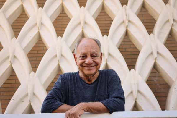 Cheech Marin smiles warmly while leaning over a white railing, standing in front of the geometric sculptural facade of The Cheech Marin Center for Chicano Art & Culture in Riverside, California. He wears a dark blue shirt. Photo by Carlos Puma, courtesy of Riverside Art Museum, 2022.