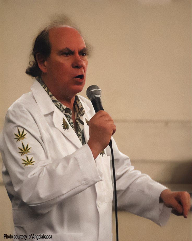 Photo of Ed Rosenthal speaking into a microphone while wearing a white coat with cannabis leaf patches, photo courtesy of Angelabacca.
